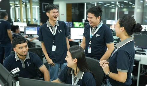Five diverse office professionals in navy polo shirts smiling and talking together around a computer workstation in a modern open-plan office.